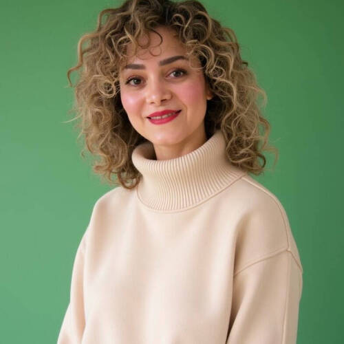 Dr. Reihaneh Hariri smiles at the camera against a green background. She has curly light brown hair and is wearing a light beige turtleneck sweater.